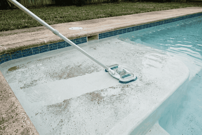 A manual pool vacuum moving over settled grey sediment on the pool floor